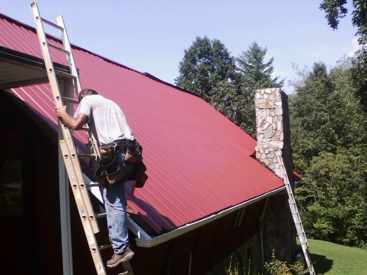 Roofer on a ladder beside a red metal roof and stone chimney — Soffit & Fascia Repair by Roofing Brothers in Patrick